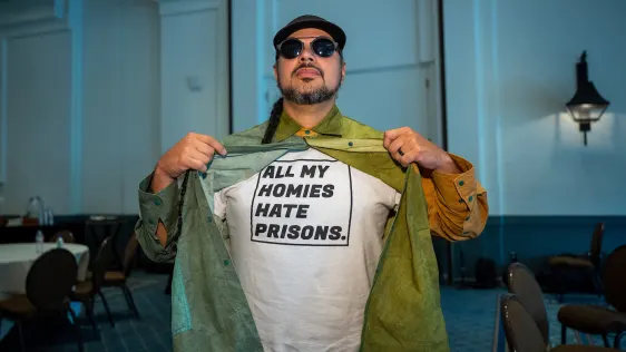 Right of Return Fellow Victor “Marka27” Quiñonez, bearded, with dark glasses and a dark hat. He holds up his unbuttoned green shirt to show the white t-shirt underneath that says, "All my homies hate prisons." [Photo by Maurice Sartirana, courtesy of Right of Return Fellowship]