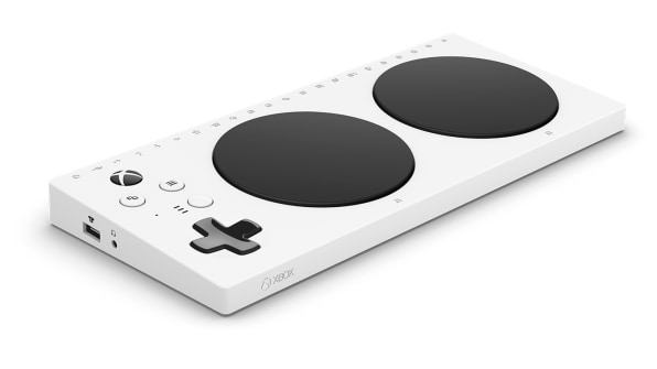A photograph of the Adaptive controller. It's a white plastic device approximately the same dimensions as a 60% keyboard, with two large touch sensitive black circles on the top. On the left hand top side of device there is a directional pad and four function keys, on the left side of the device a headphone jack and usb port are visible.