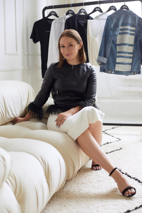 A portrait of Aille Design founder Alexa Jovanovic, seated on a modern sofa in front of a rack of clothes.